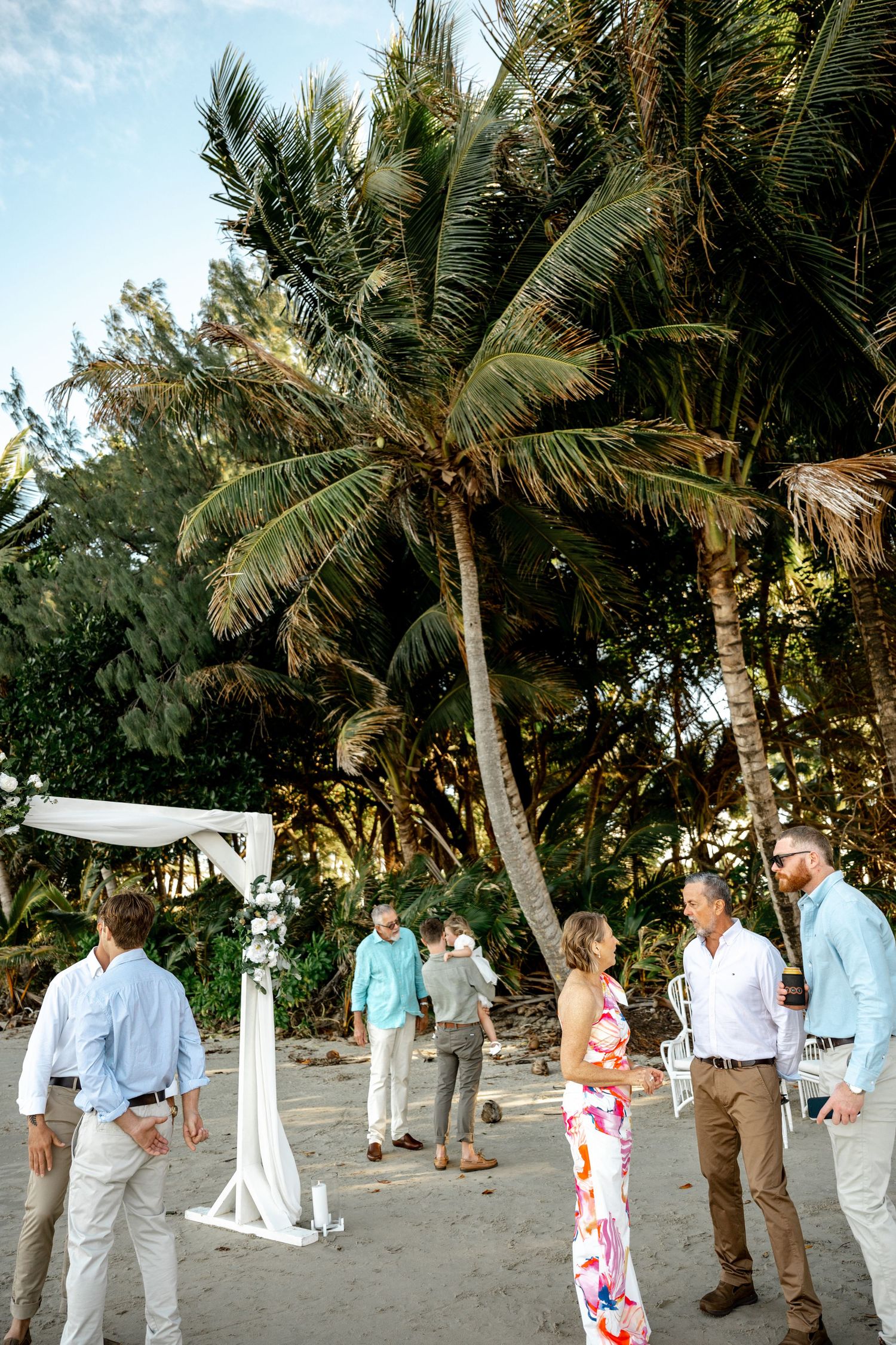 Wedding guests mingle near a white floral arch on a tropical beach surrounded by tall palm trees at Four Mile Beach, Port Douglas.