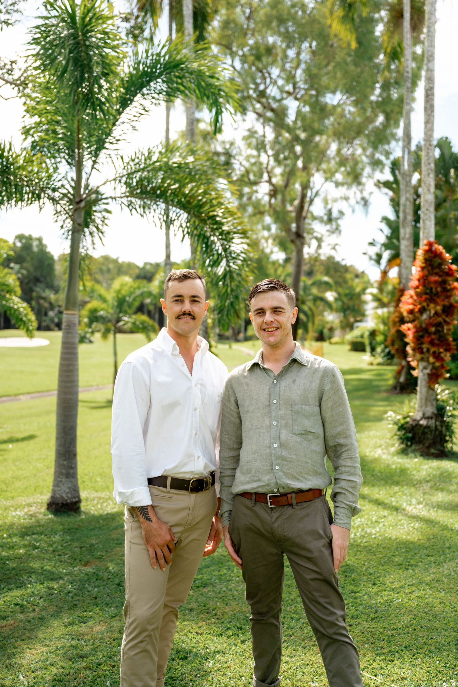 Two men in smart casual shirts posing together in a tropical garden with palm trees behind them.