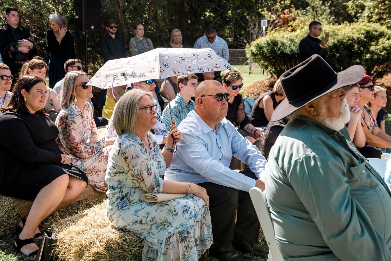 Wedding guests seated outdoors, one holding a floral umbrella for shade, man in hat visible in foreground.