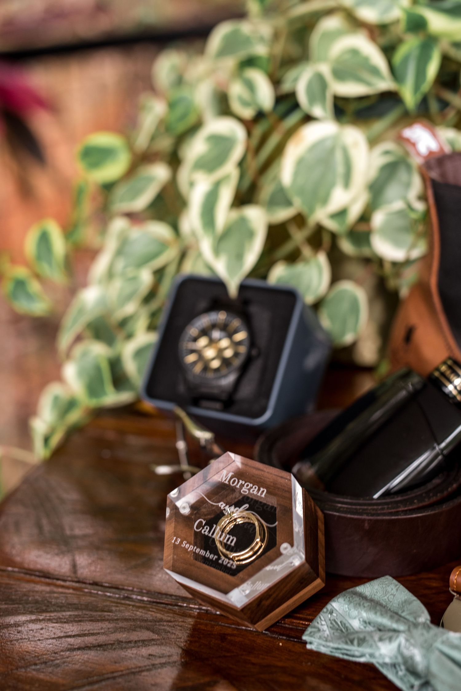 Custom engraved hexagonal acrylic ring box with wedding rings beside a leather belt and watch on wooden table.