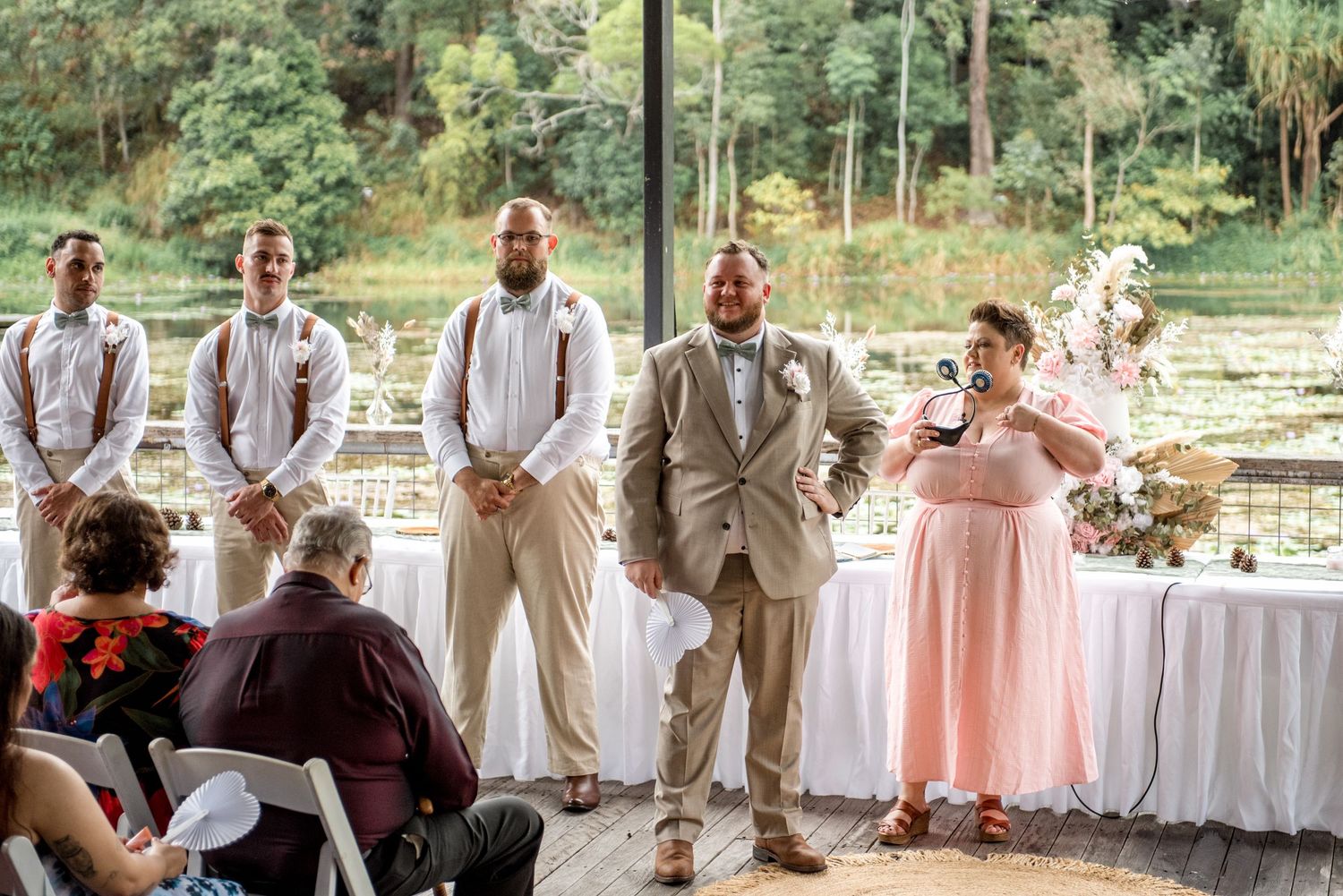 Groom in tan suit standing with groomsmen in suspenders during outdoor wedding ceremony.