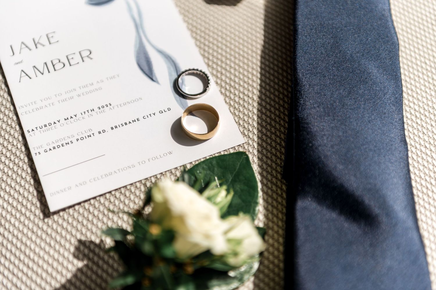 Wedding flat lay featuring Jake and Amber invitation, gold rings, navy tie and boutonniere.