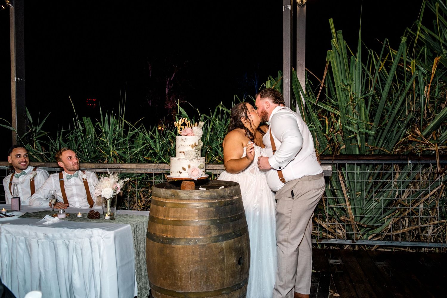 Bride and groom share a kiss while cutting their wedding cake on a rustic barrel outdoors.