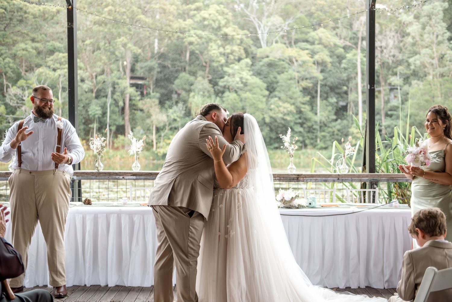 Bride and groom share first kiss at wedding ceremony with green forest backdrop and wedding party watching.