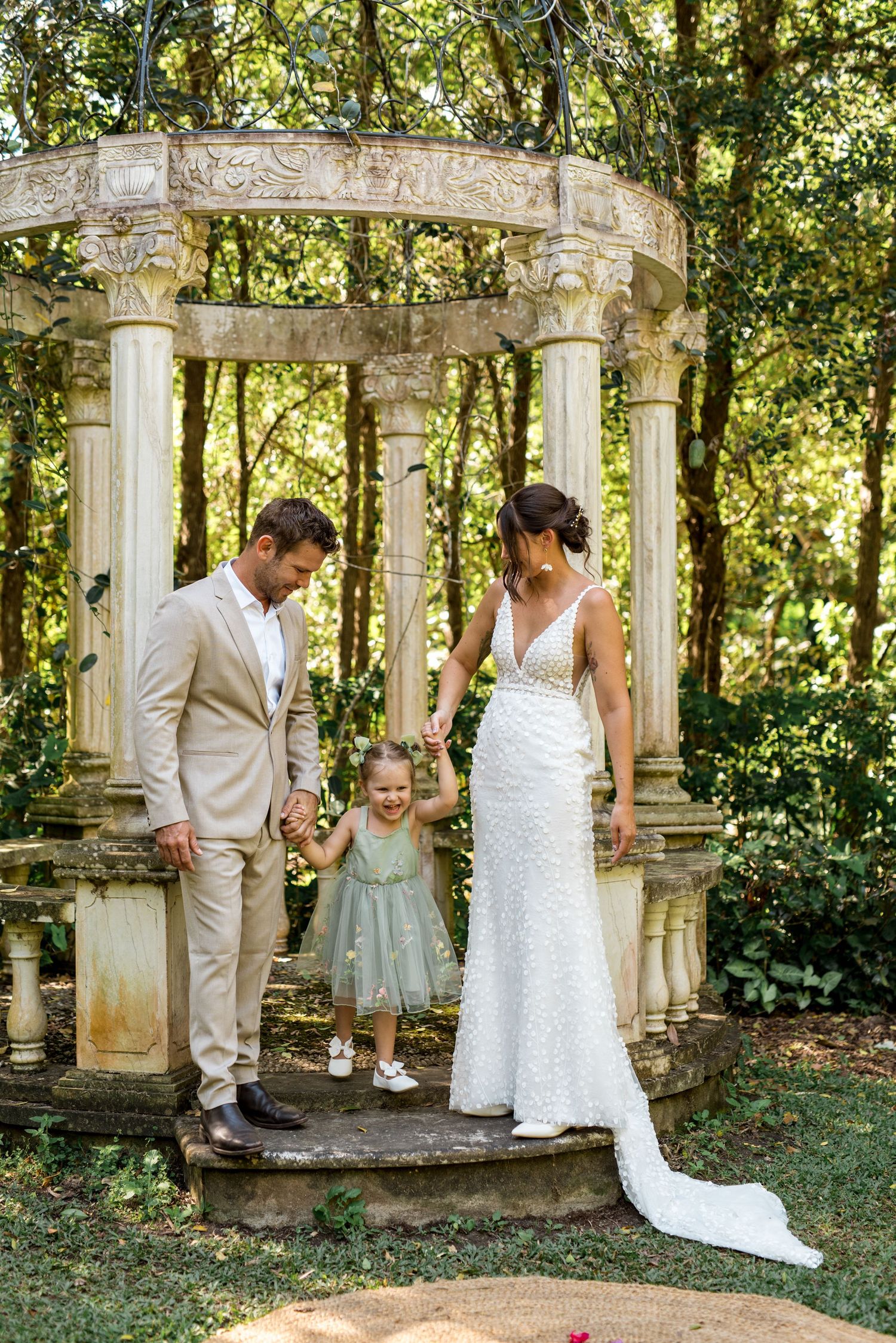 Bride and groom lifting smiling daughter between them inside ornate stone rotunda at outdoor garden wedding.