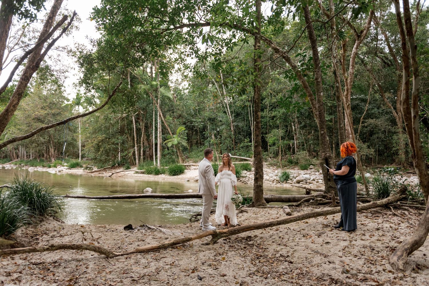 Officiant leads intimate elopement ceremony for couple on sandy riverbank in tropical forest.