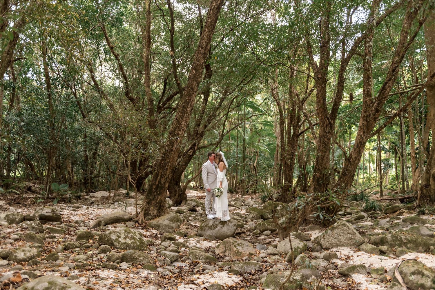 Bride and groom standing together among rocks in a serene lush forest setting.