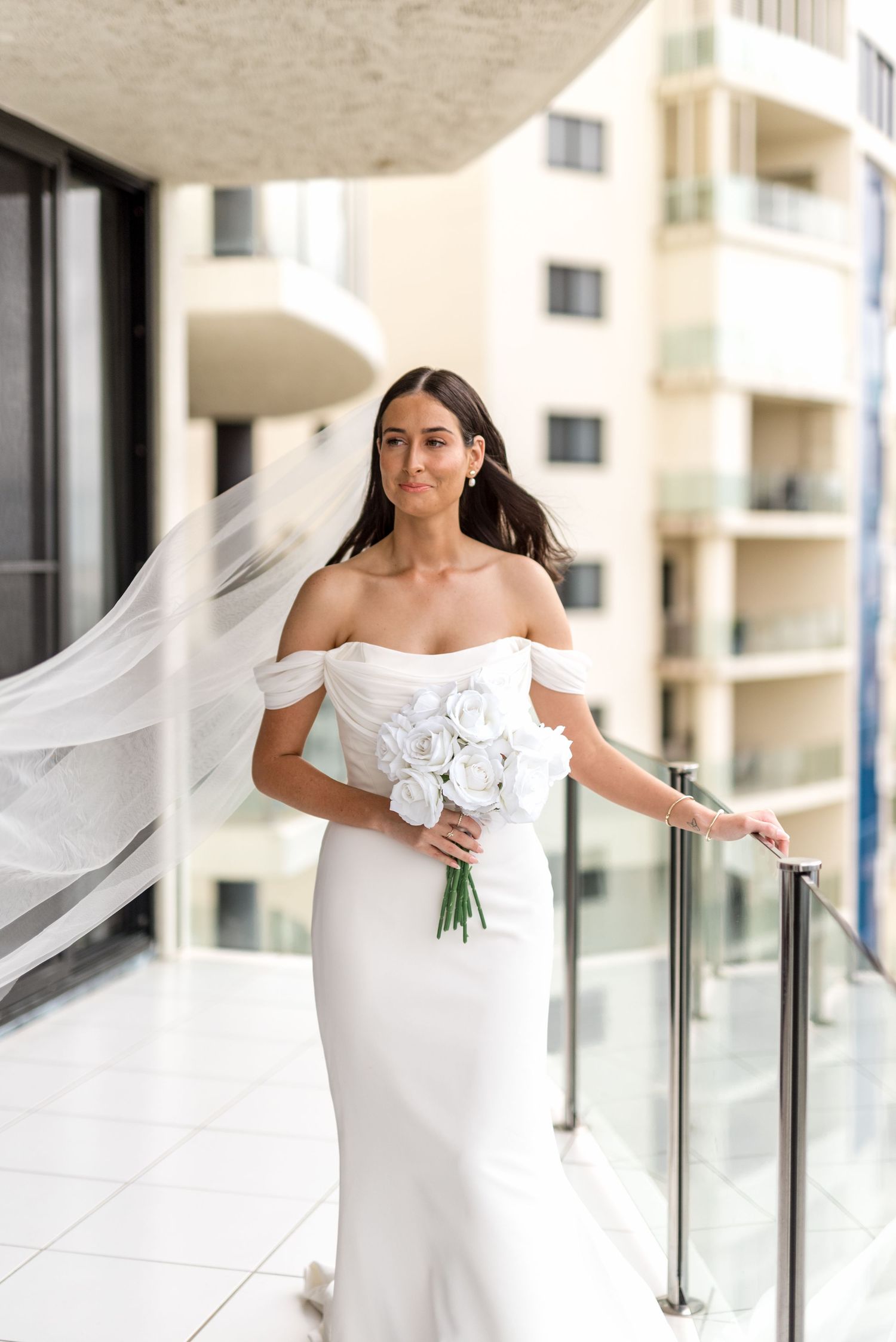Bride in white off-shoulder gown with flowing veil holding white roses on balcony.