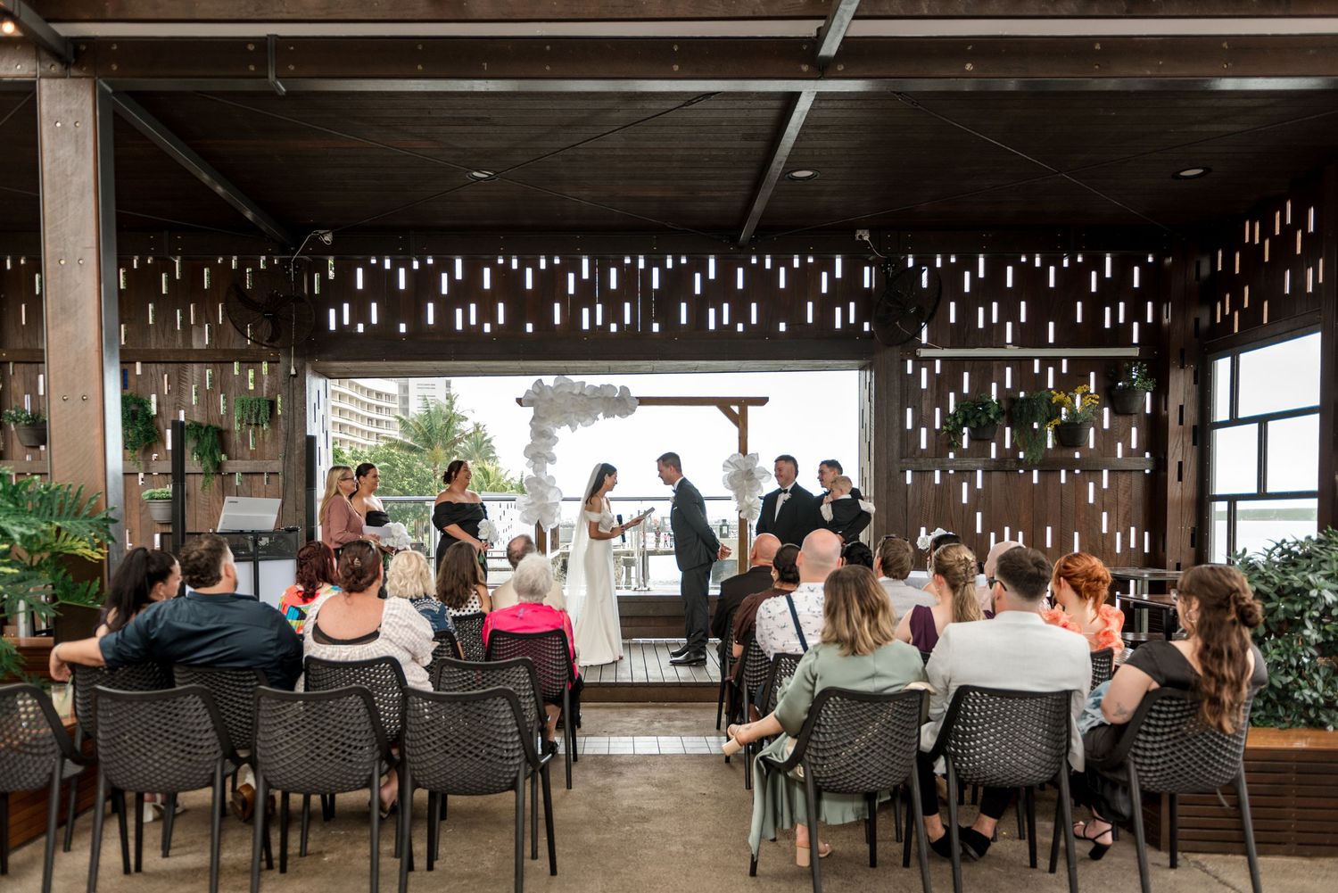 Small wedding ceremony with seated guests facing couple at altar with decorative hanging lights.