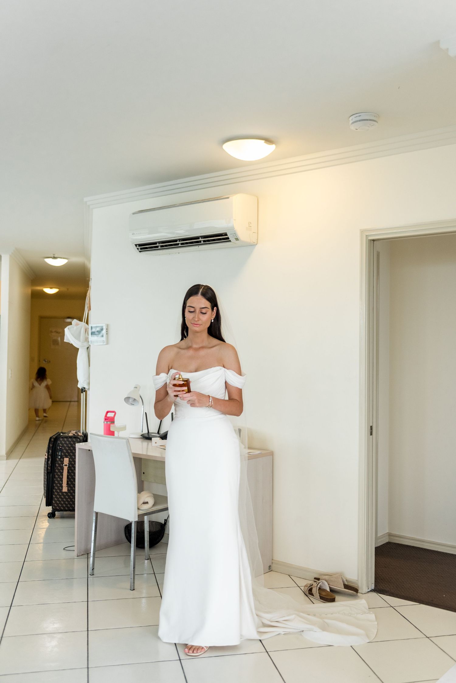 Elegant bride in white off-shoulder wedding gown checks phone while standing in bright hotel room.