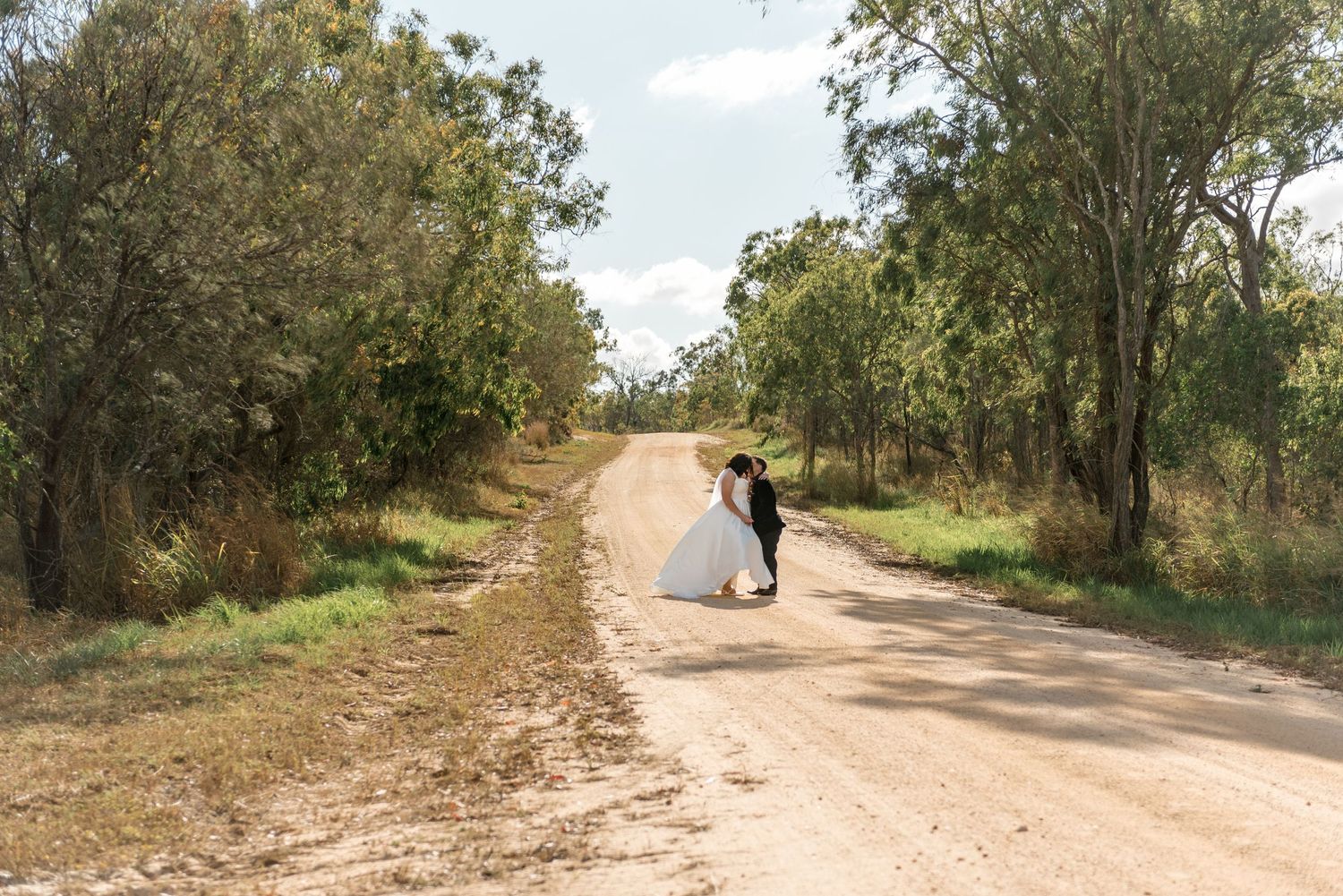 Bride and groom share a romantic dip kiss on a sandy dirt road surrounded by lush green trees.