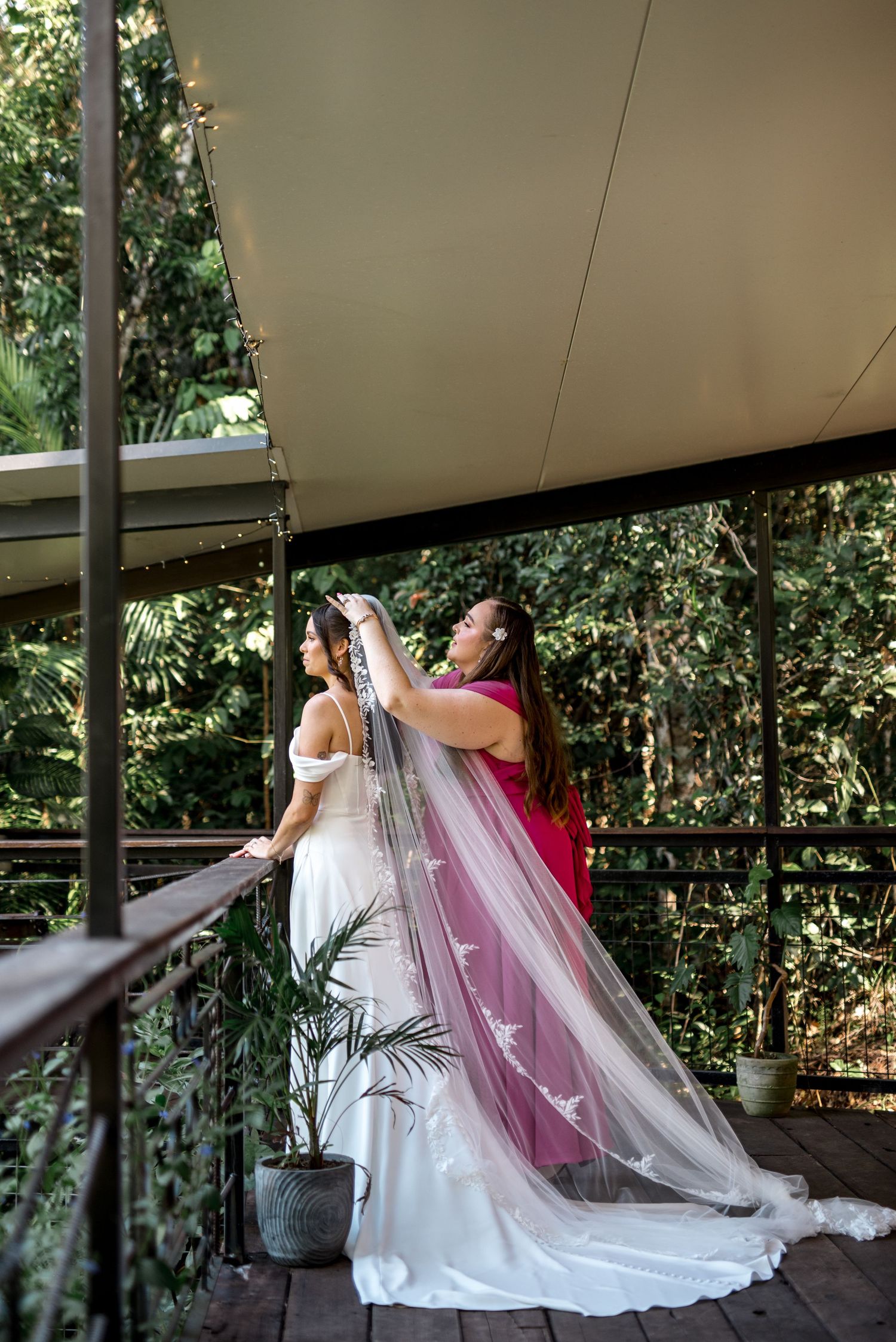 Bridesmaid in pink dress arranging bride's long embroidered veil on a balcony overlooking tropical greenery.