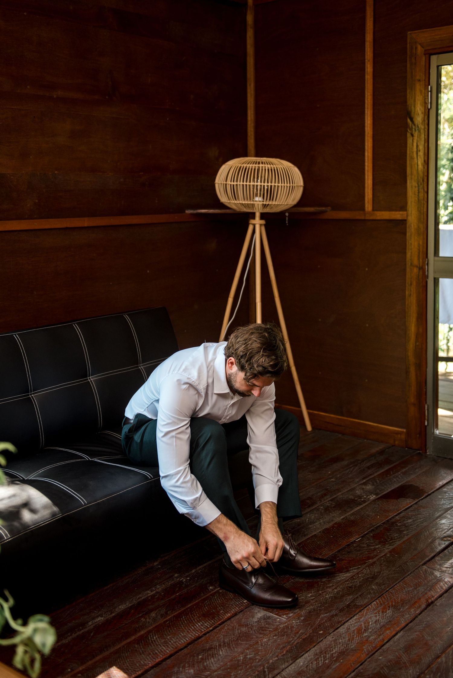 Groom in white shirt and dark trousers putting on brown leather dress shoes on a wooden floor.