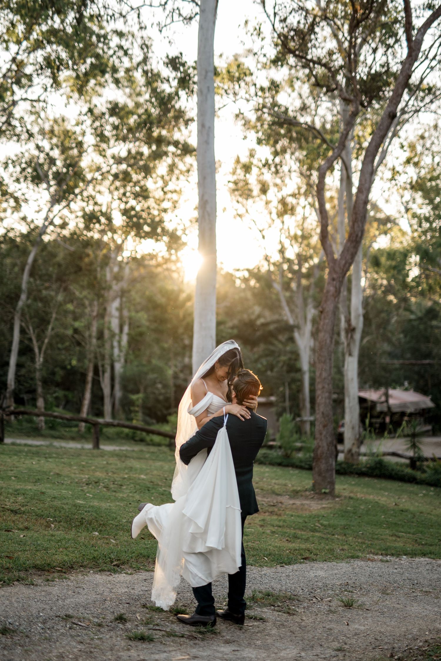 Groom lifts bride joyfully in a sunlit forest clearing during wedding portraits.