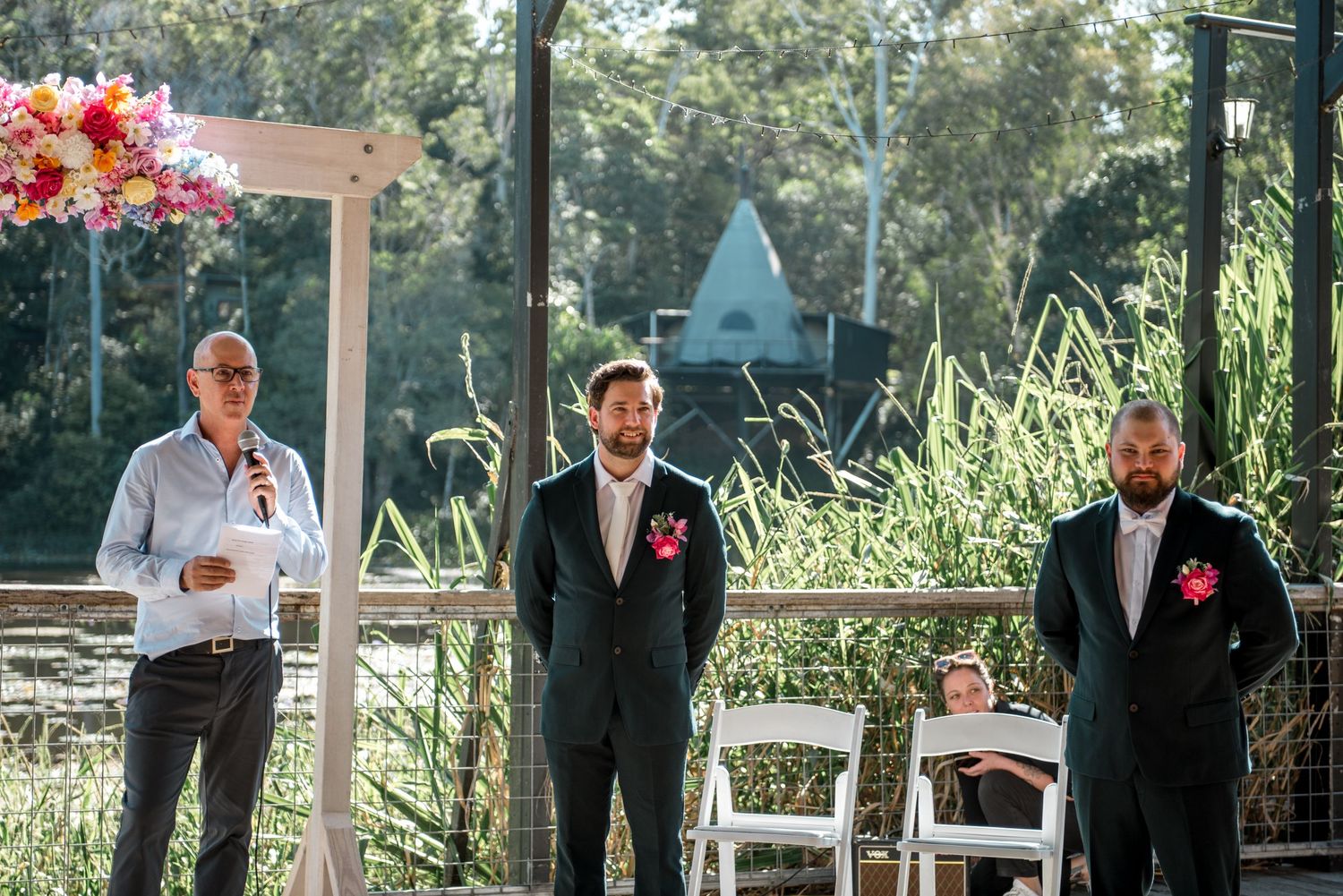 Groom and best man stand at outdoor wedding ceremony altar decorated with colorful pink flowers.