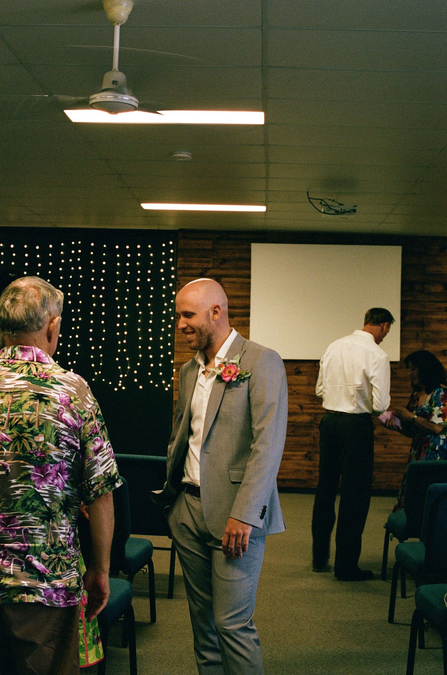 Smiling bald groom in grey suit with pink boutonniere greeting guests at ceremony.