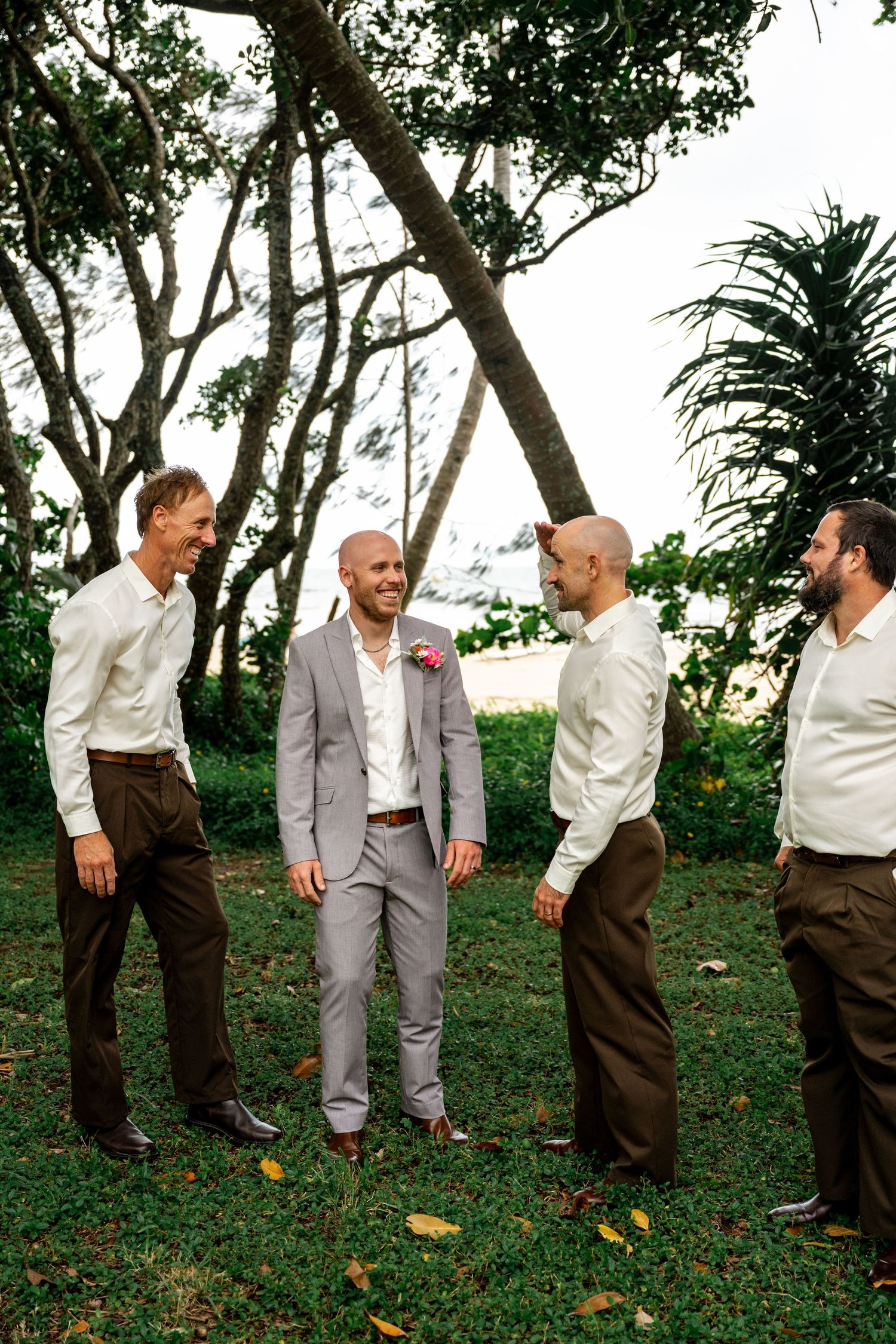 Groom in grey suit laughs with three groomsmen in white shirts and brown pants under tropical trees at outdoor wedding.