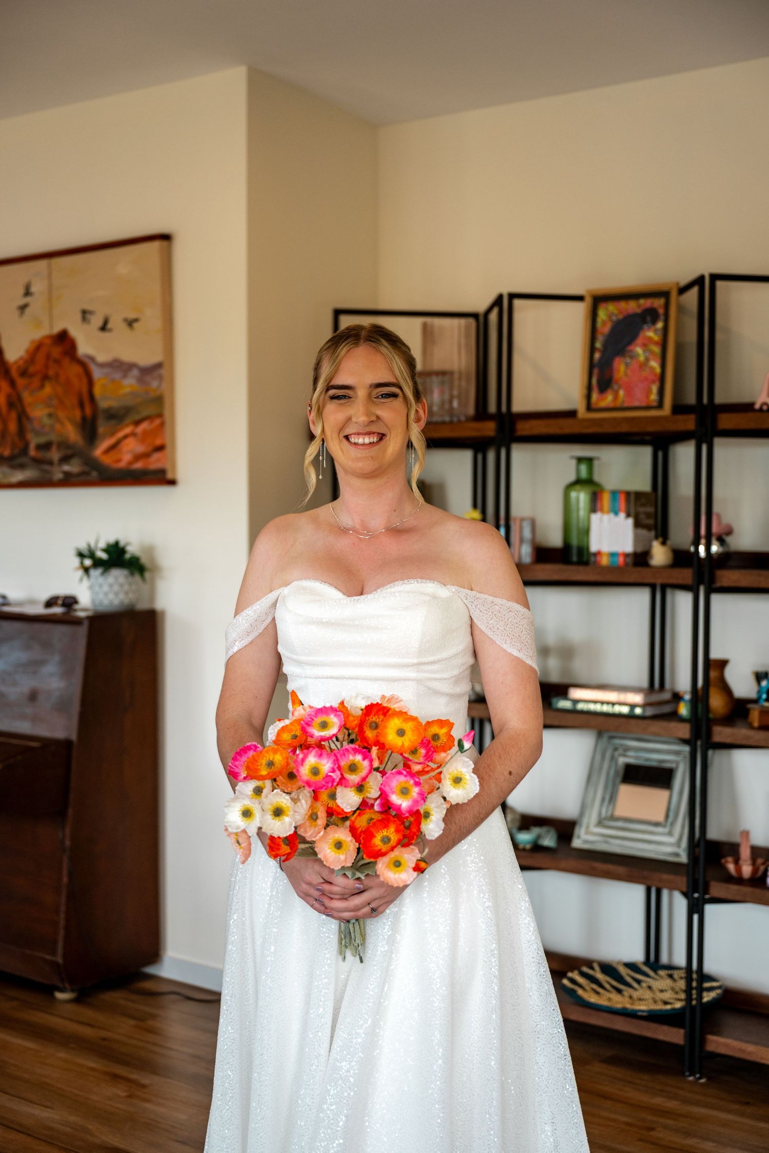 Smiling bride in white off-shoulder wedding dress holding colorful orange and pink bouquet indoors.