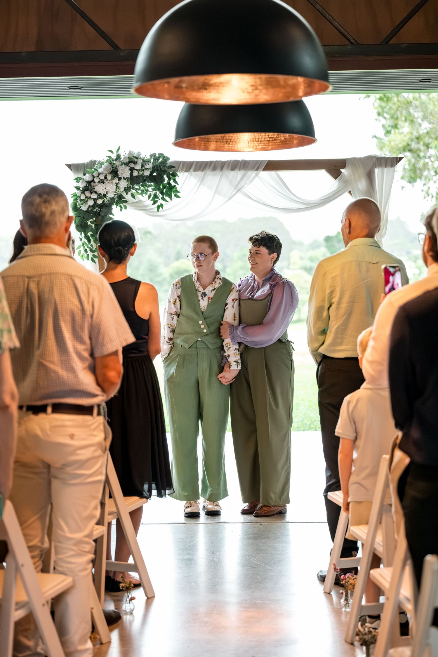 Two brides in green suits standing at altar during wedding ceremony viewed from aisle with pendant light above.