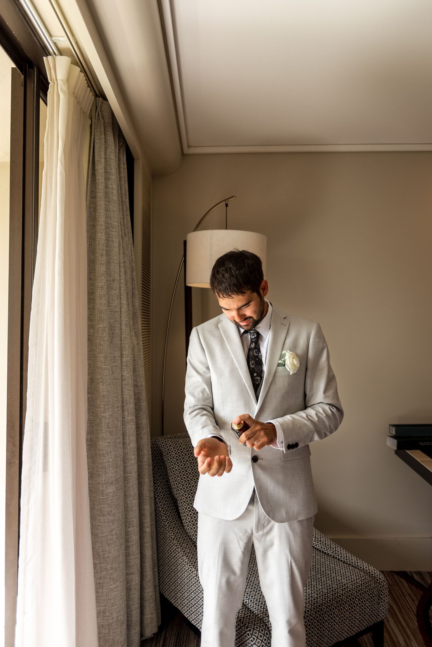 Groom in elegant light gray suit with dark floral tie fastening his cufflinks by a window in a hotel room.