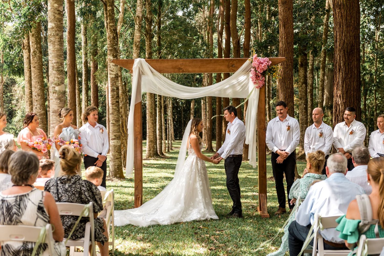 Bride and groom standing at a floral wooden arch altar during an outdoor forest wedding.