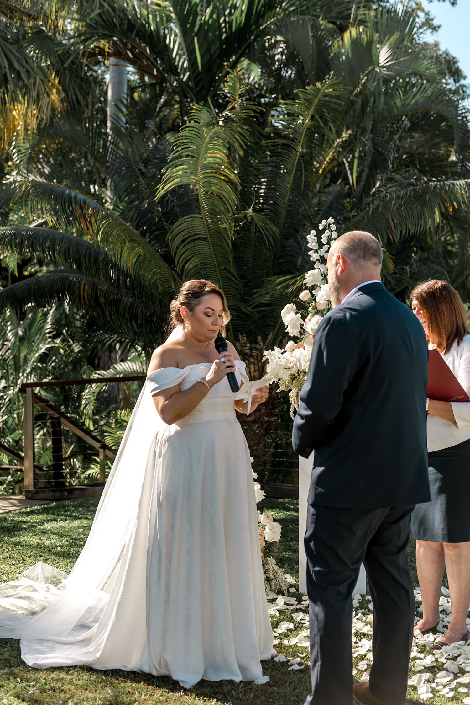 Bride in off-shoulder gown reads vows into microphone at outdoor tropical wedding ceremony with palm trees.