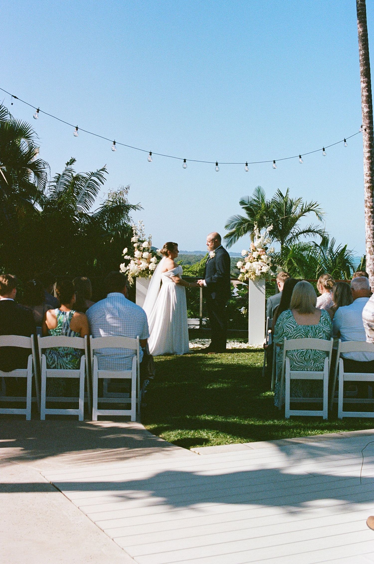 Outdoor wedding ceremony on a sunny lawn with white chairs, tropical palms, and ocean views.