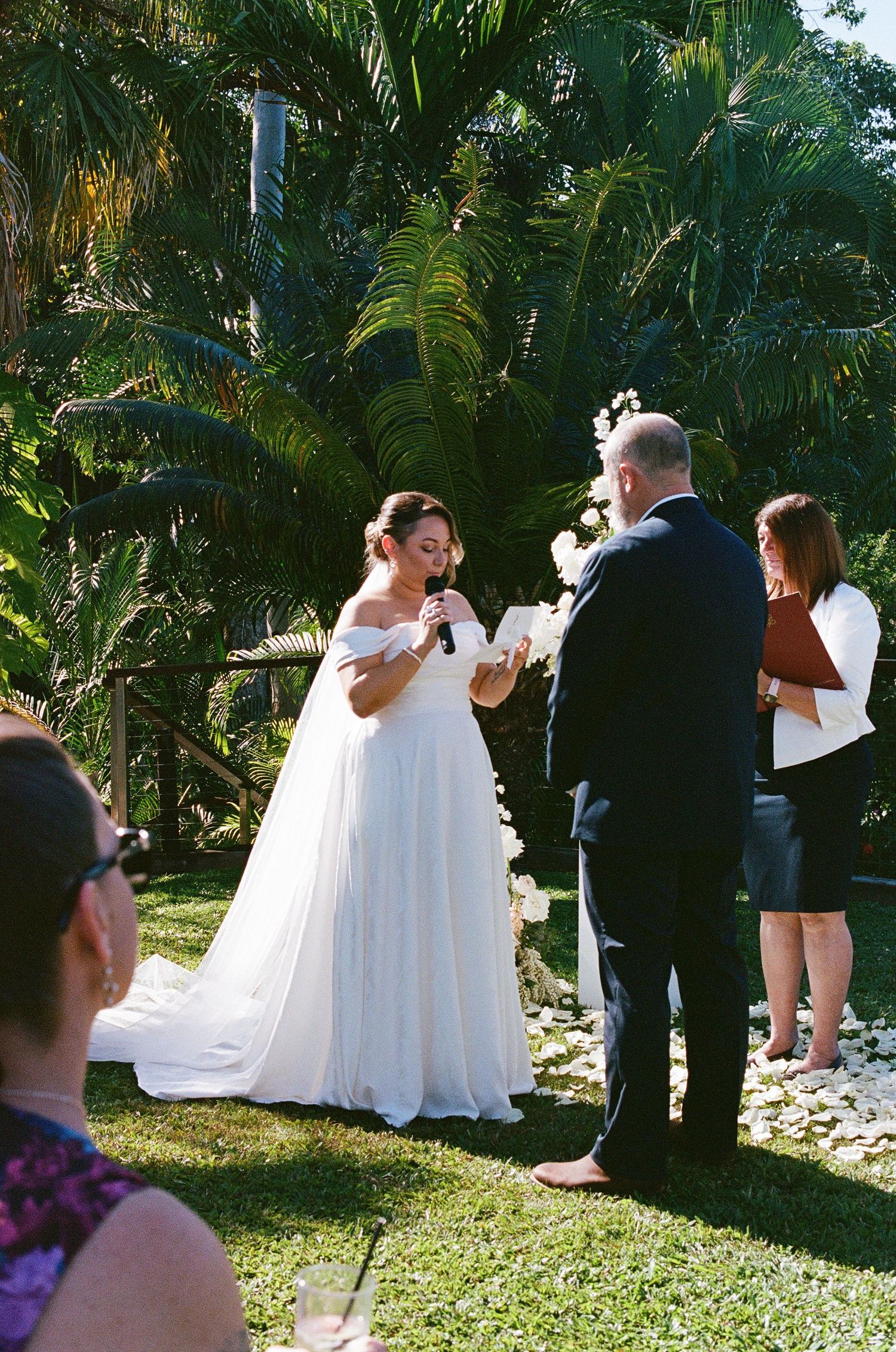 Bride in off-shoulder white gown reading vows outdoors surrounded by tropical greenery.