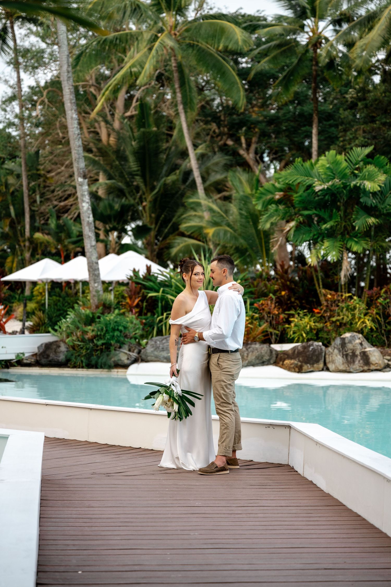 Bride and groom standing together on a wooden deck beside a tropical resort lagoon pool at Sheraton Grand Mirage Port Douglas.