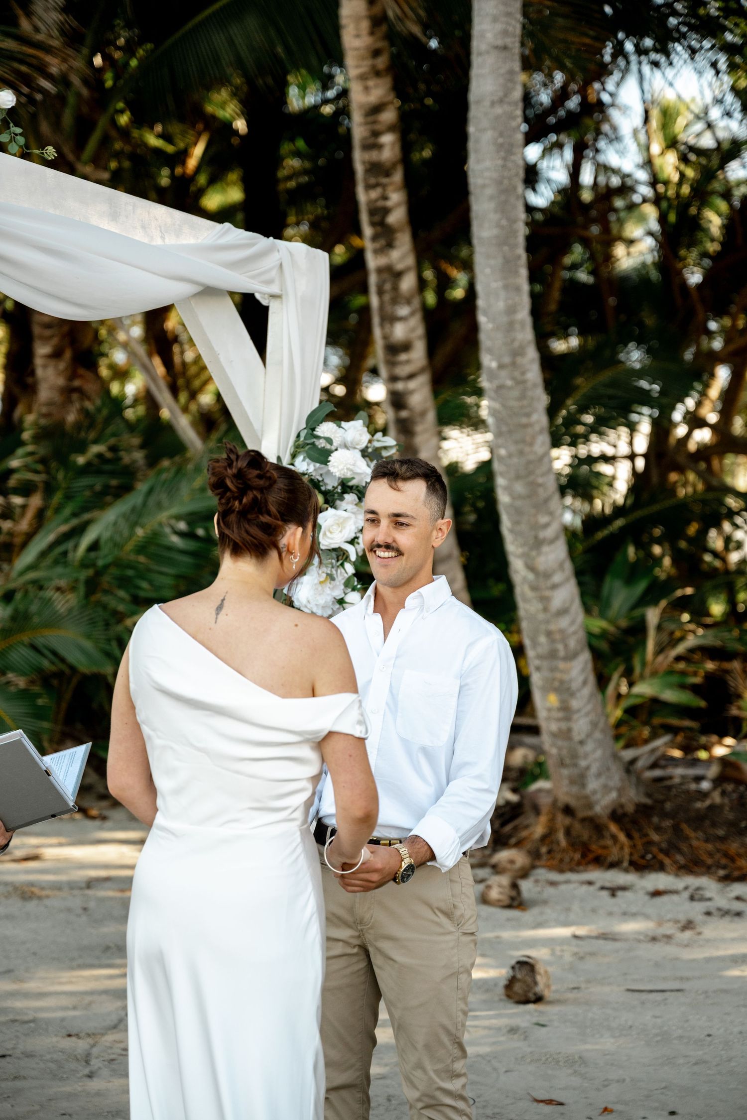 Groom smiling lovingly at bride during beach wedding ceremony under white floral arch with palm trees surrounding at Four Mile Beach.