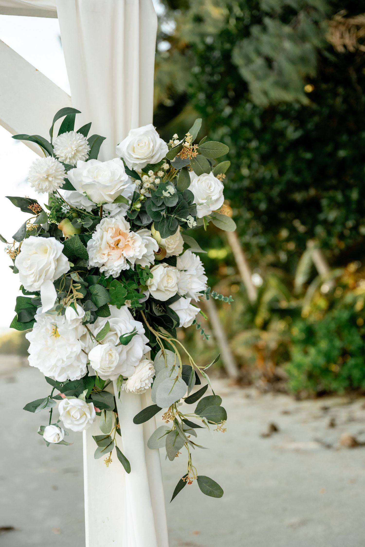 Elegant wedding arch decorated with white roses, peonies, and eucalyptus greenery on a beach at Four Mile Beach, Port Douglas.