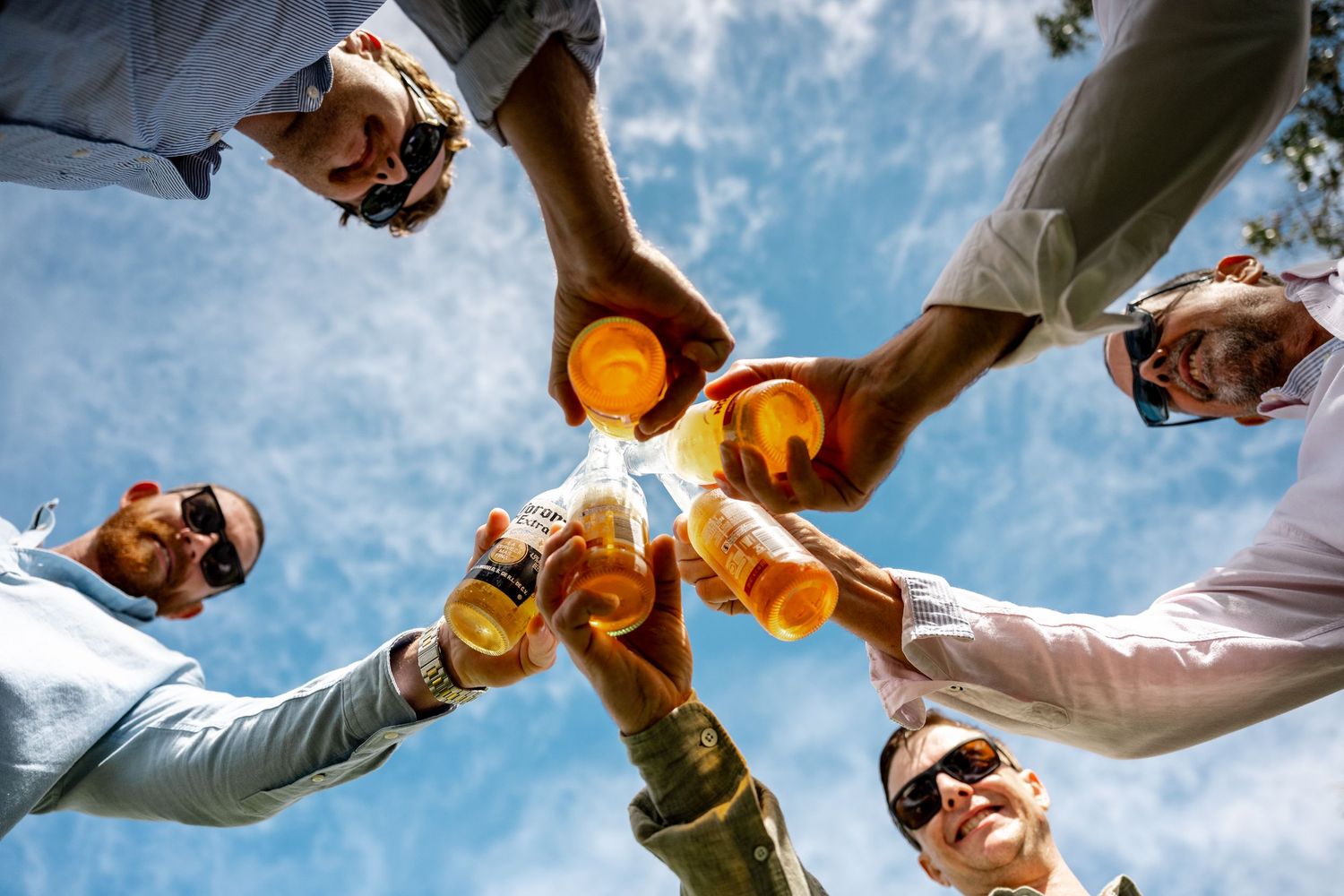 Low angle view of five men clinking beer bottles together against a bright blue sky.