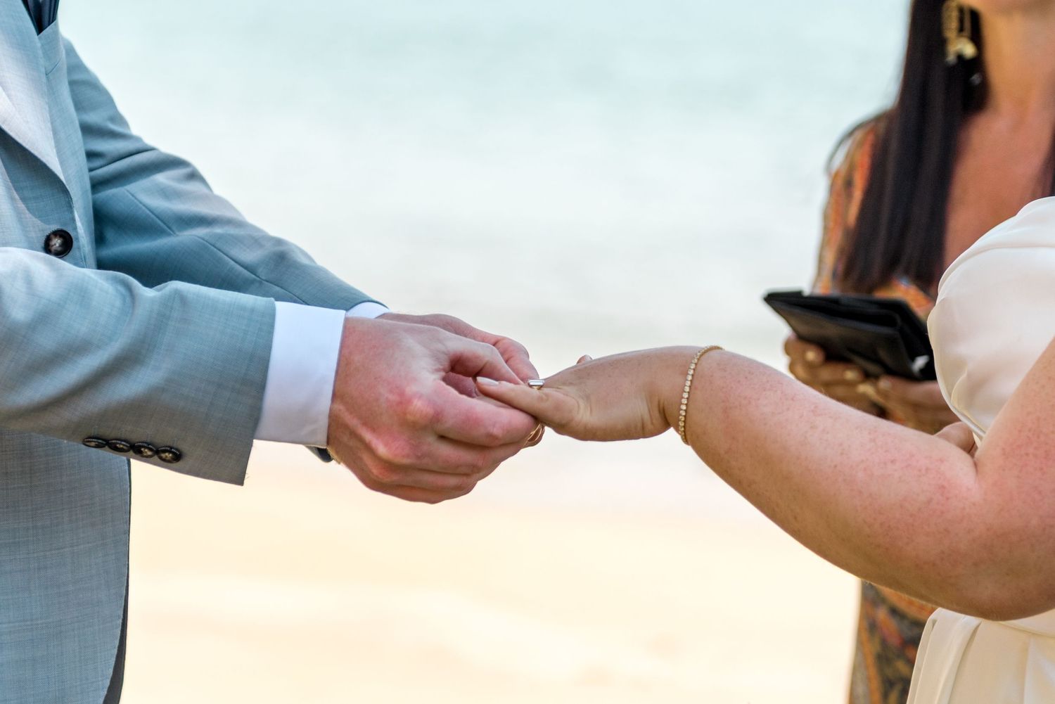 Groom placing wedding ring on bride's finger during beach ceremony.