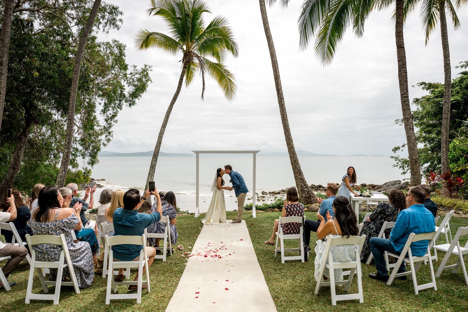 Intimate beach wedding ceremony with guests seated on white chairs under swaying palm trees at Little Cove, Port Douglas.