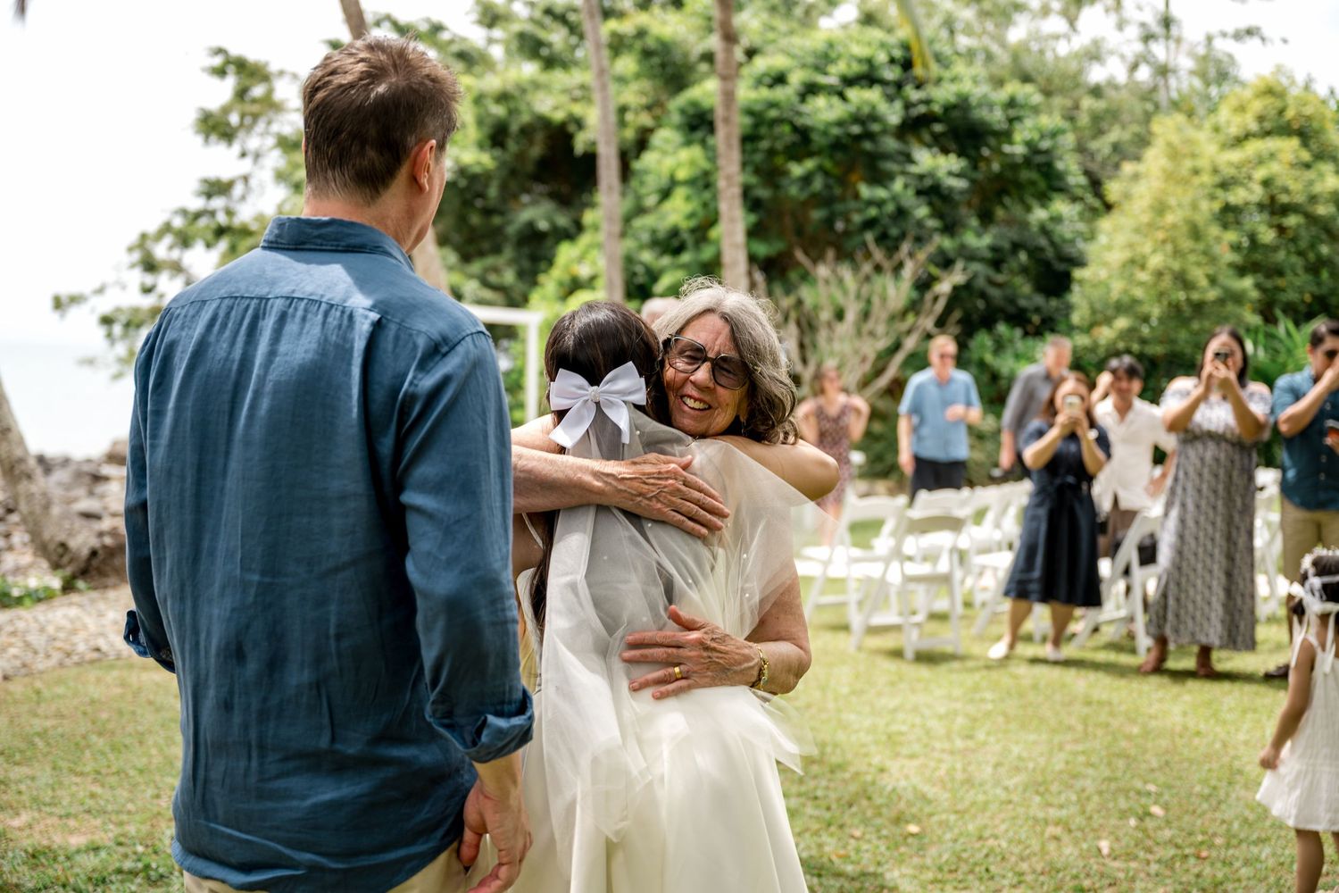 Bride hugging a guest at an outdoor tropical wedding ceremony while groom stands nearby at Little Cove, Port Douglas.