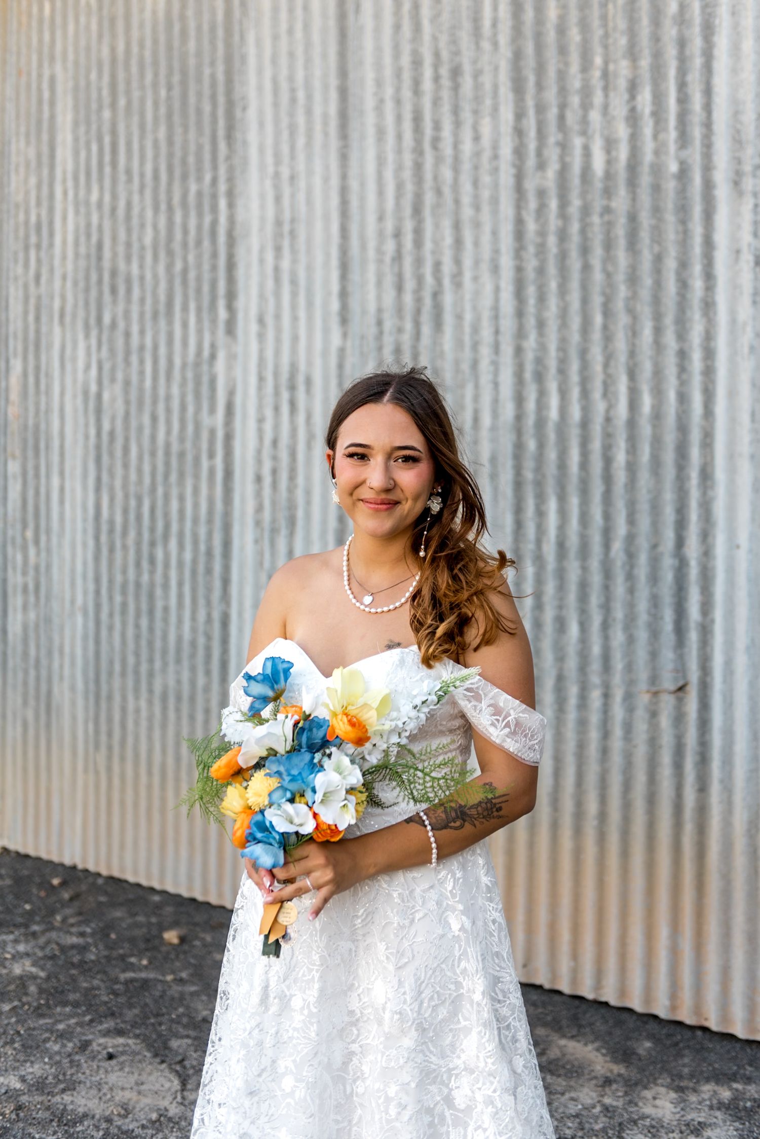 Smiling bride in lace gown holds colorful blue, white, and orange bouquet against corrugated metal wall.