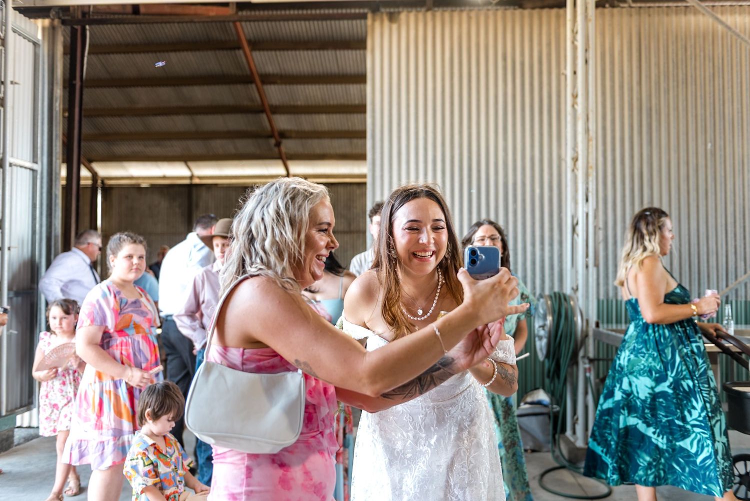 Bride in white lace gown takes a selfie with a guest in a pink dress at a barn wedding reception.