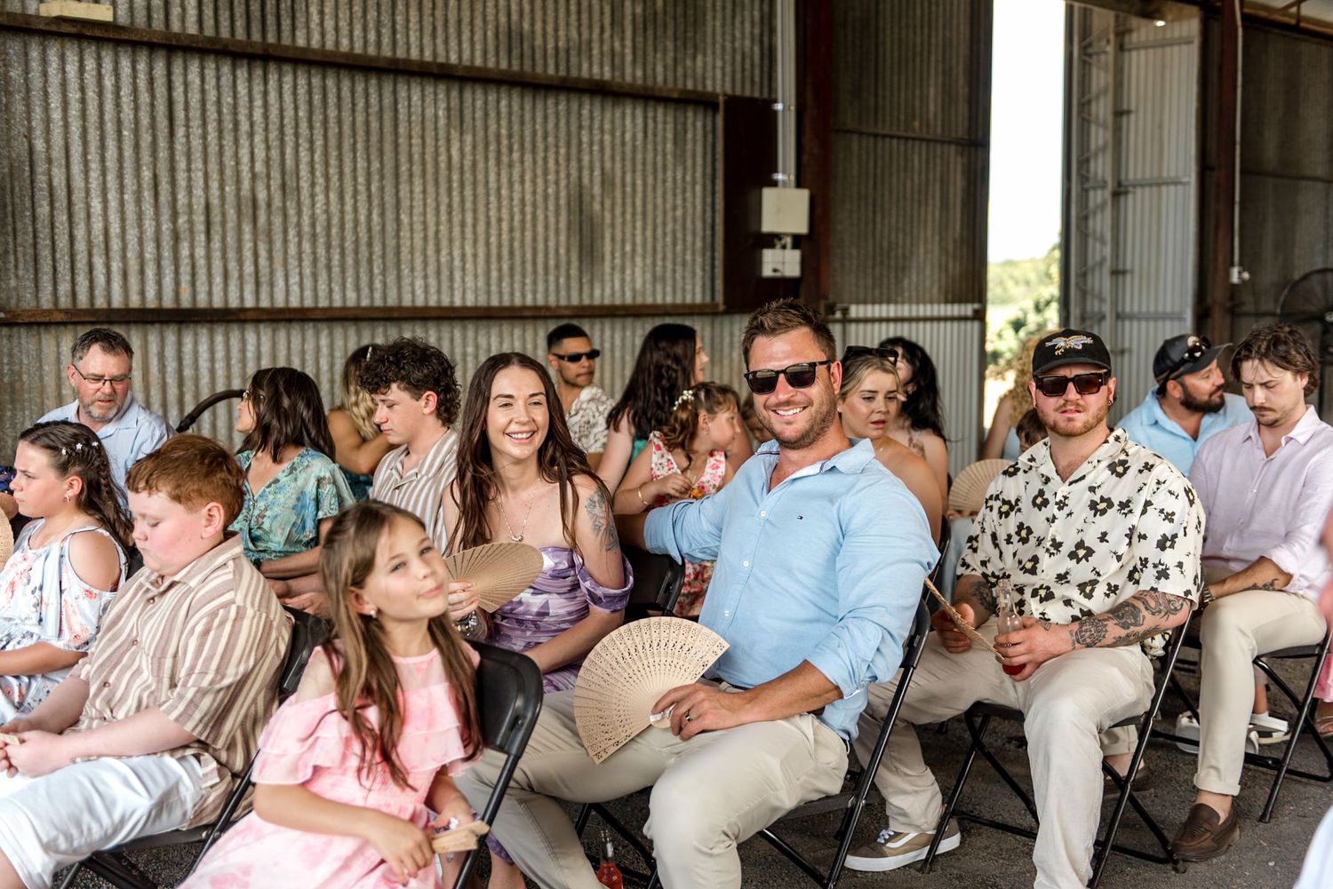 Smiling wedding guests in summer attire seated in a barn, with a man in a blue shirt holding a fan.