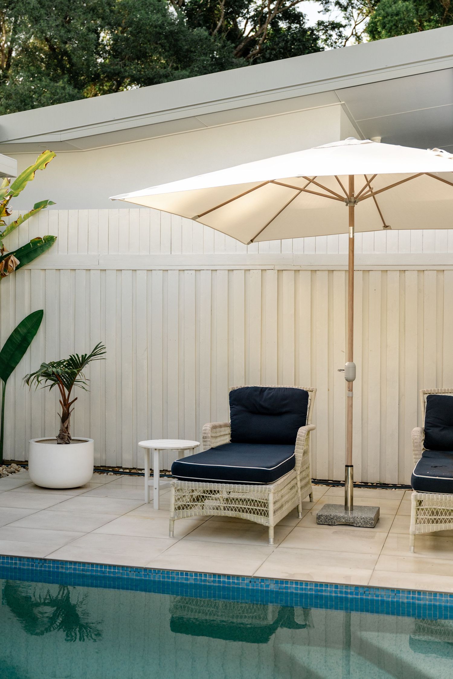 Luxury pool area with white wicker lounge chairs, navy cushions, and white umbrella beside fence.