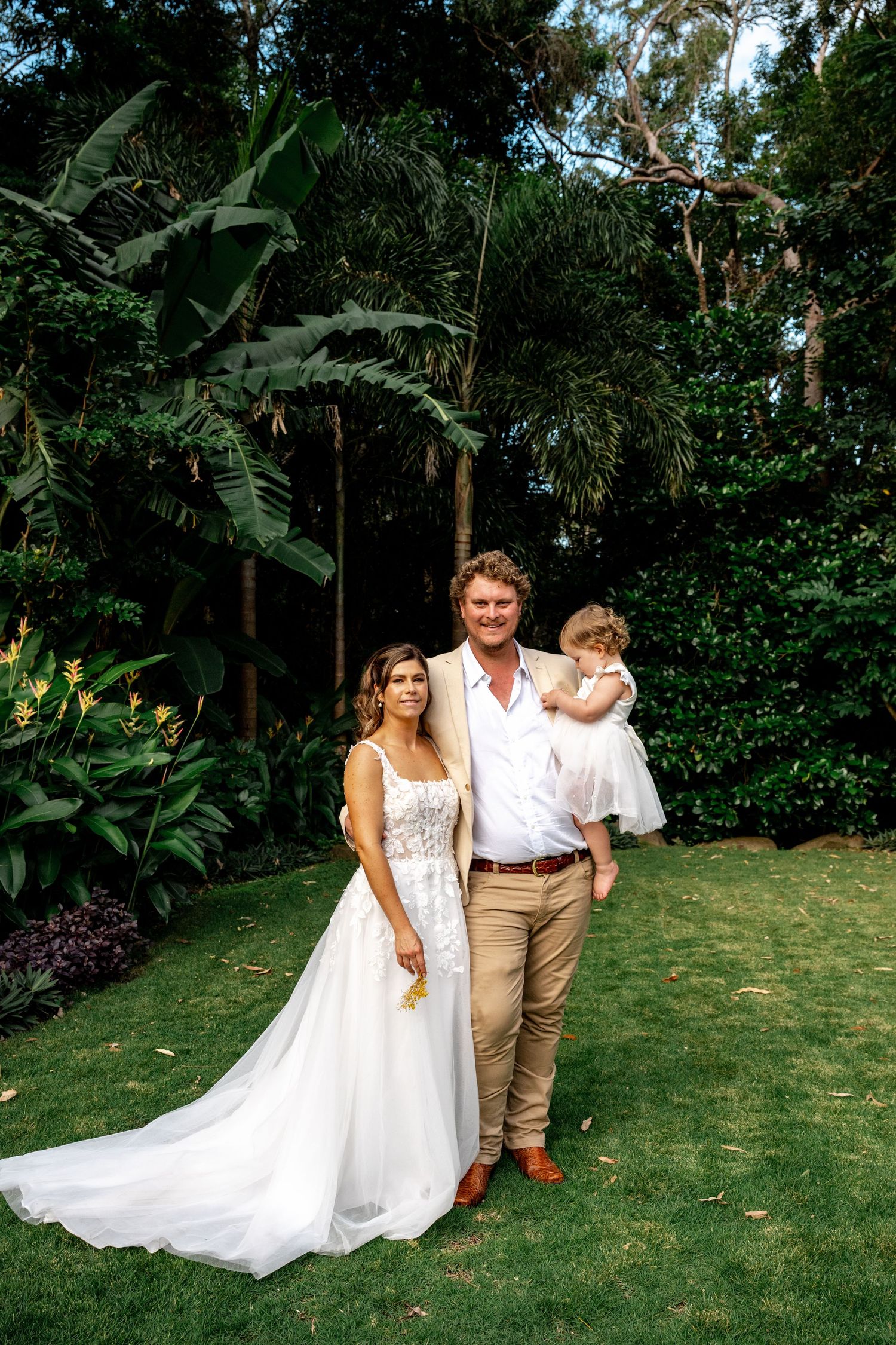Bride and groom posing with young child in white dress in tropical garden.