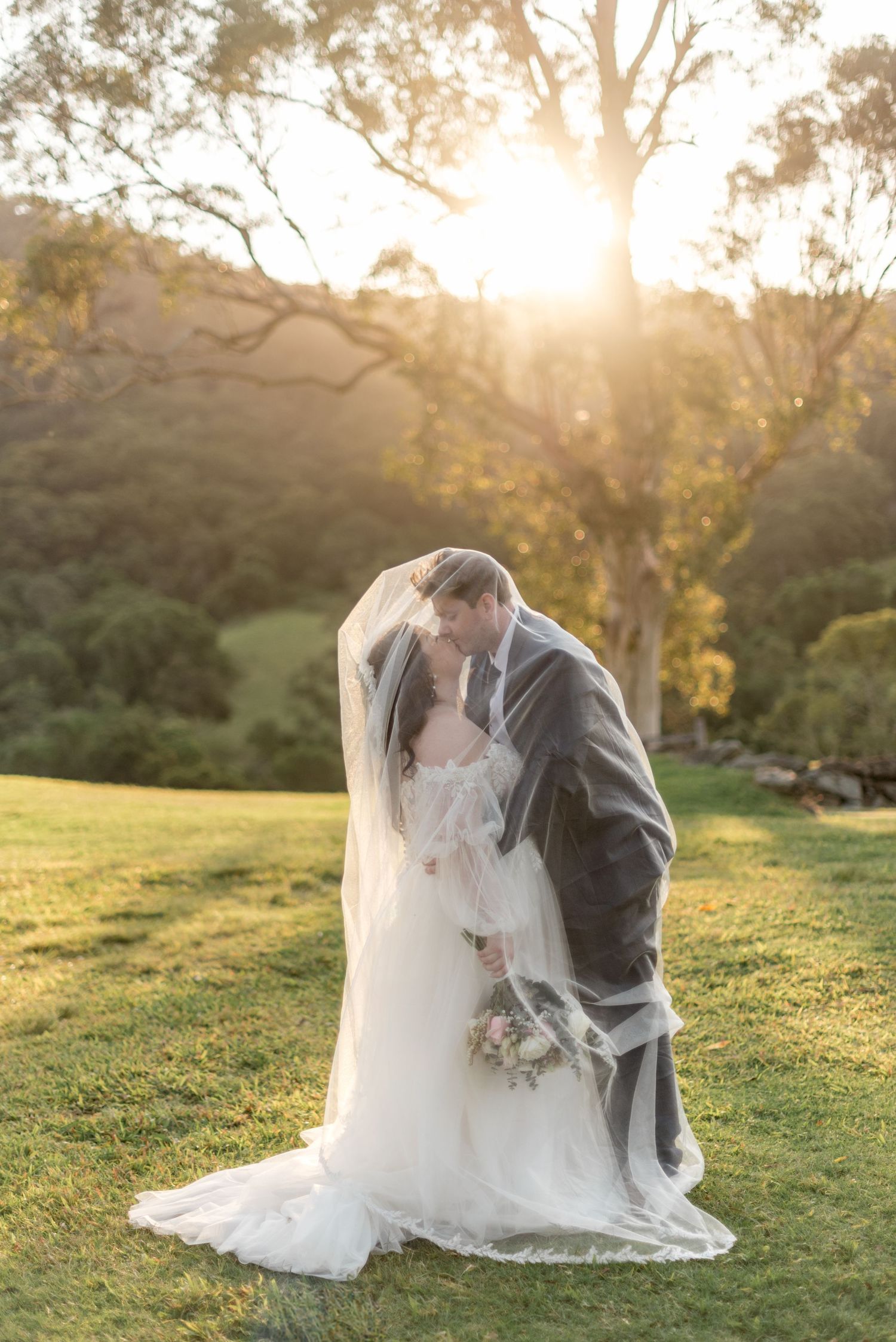 Bride and groom kissing under flowing veil with golden sunlight through trees behind them.