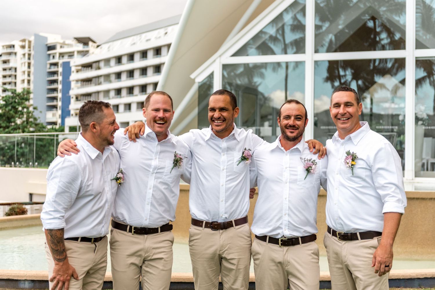 Five groomsmen in white shirts and khaki pants laughing together at a tropical wedding venue.