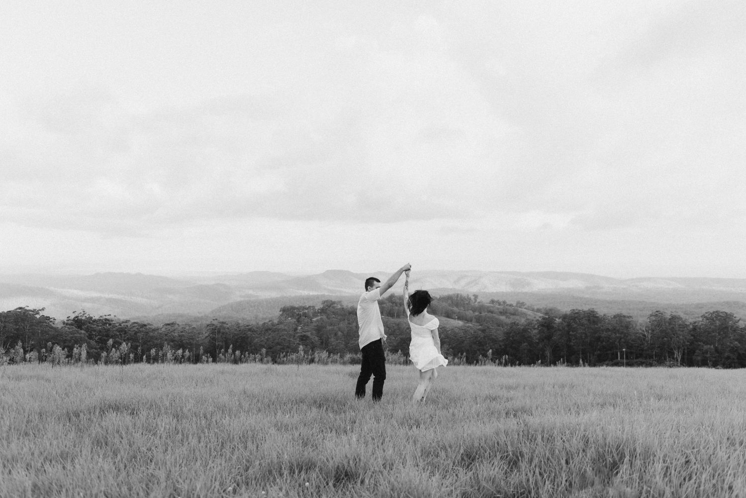 Black and white photo of couple dancing in open field with scenic mountain landscape behind.