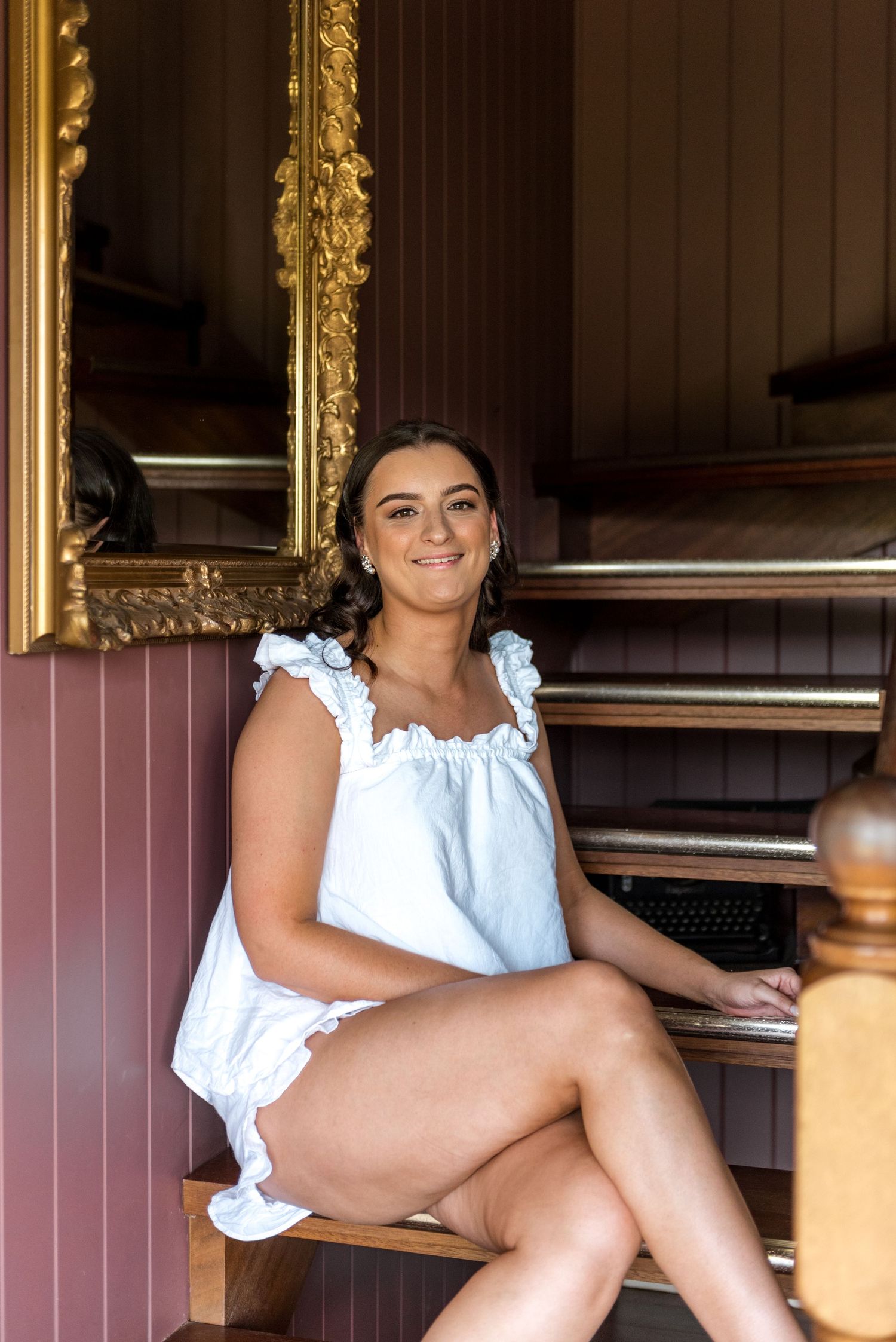 Smiling bride in white ruffled top sitting on staircase beside ornate gold mirror.