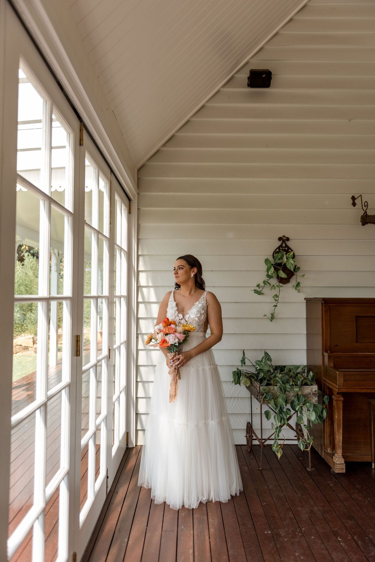 Bride in white tulle gown holding colorful bouquet gazing out window on timber verandah.