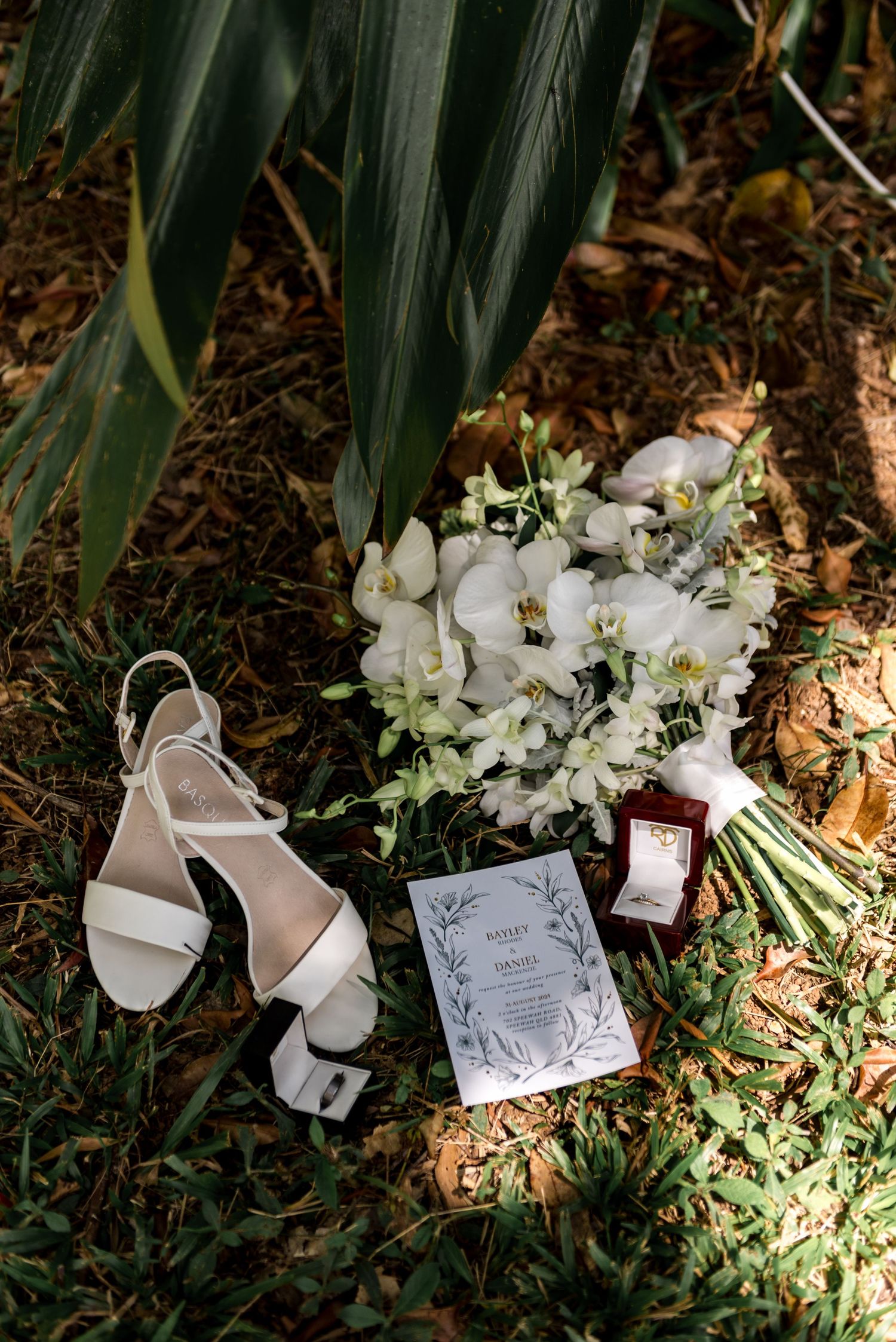 Wedding flat lay featuring white heels, orchid bouquet, invitation, and ring box on grass.