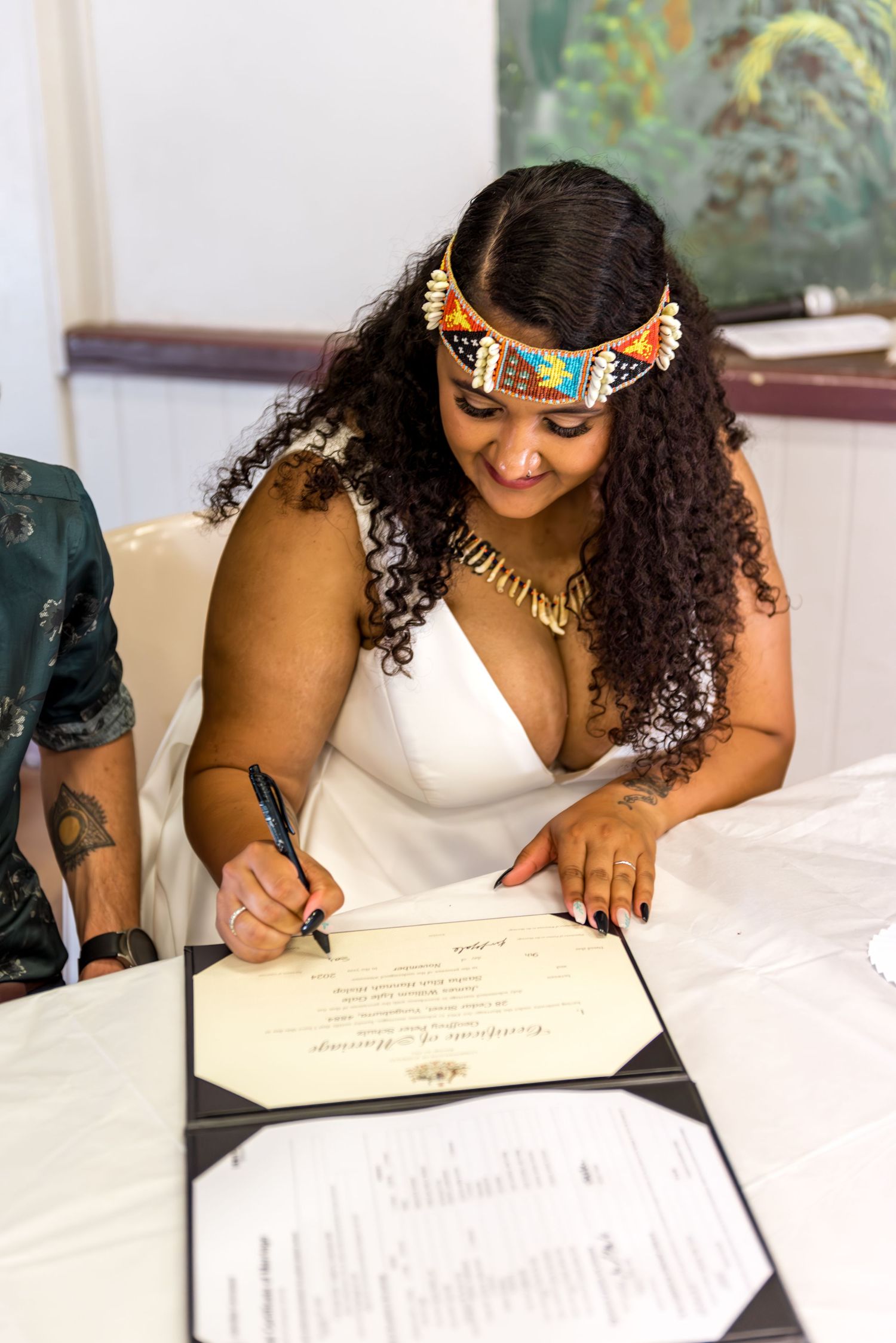 Bride with colorful beaded headpiece and gold necklace signs marriage certificate at wedding ceremony table.