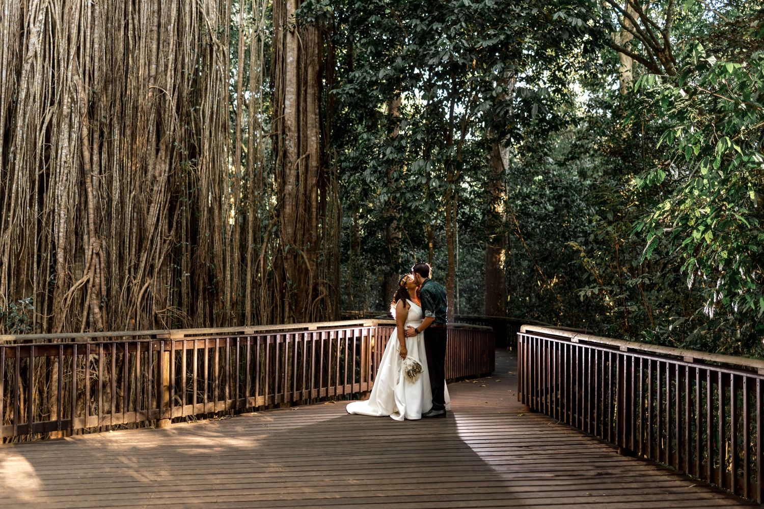 Bride and groom sharing a romantic moment on a wooden bridge surrounded by banyan trees.