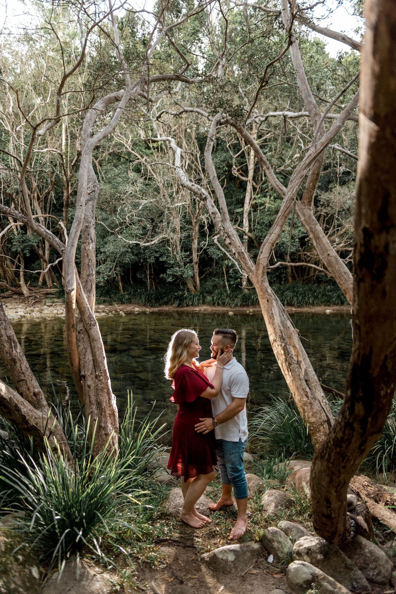 Couple holding each other tenderly by a forest waterway framed by gnarled trees and green plants.