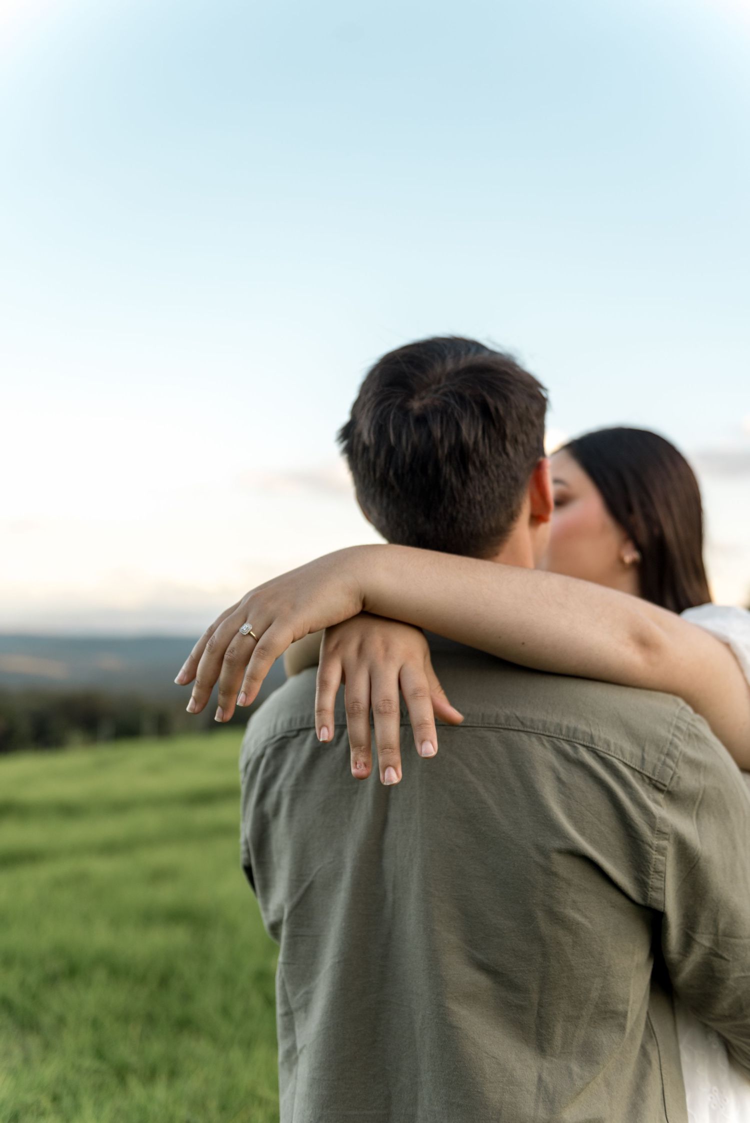 Woman in white dress kissing man in olive shirt while embracing outdoors in green meadow at dusk.